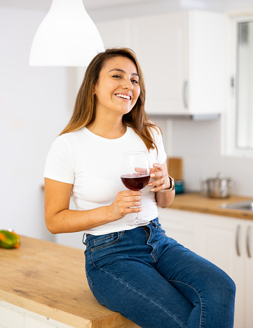 Portrait of relaxed young woman sitting on kitchen countertop, drinking red wine