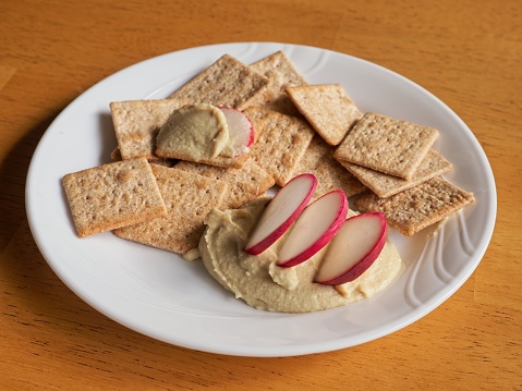 A nutritious snack of organic hummus, radishes and crackers. Red radish slices dipped in creamy humus surrounded by wheat crackers of a white plate.