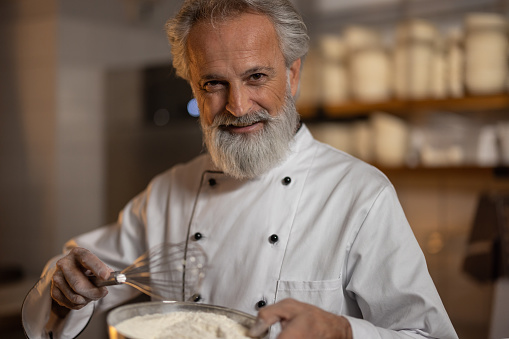A gray-haired bearded baker in a white baker's uniform prepares dough, mixes flour, eggs in the morning rays filtering through the window in the bakery. Night shift.