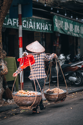 Hanoi, Vietnam - November  20, 2022: Street vendor carrying transporting goods in baskets using a carrying pole, also called a shoulder pole.