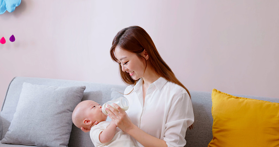 asian smiling mother is feeding little baby drinking milk from bottle and infant lying on her hand