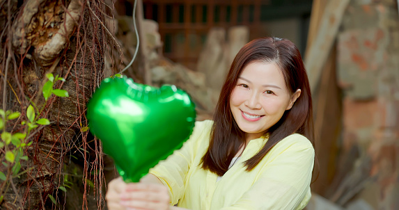 portrait Asian woman standing in front of tree showing care with mother nature - she smile hand hold green heart shape ballon