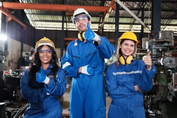 Industrial workers team in safety uniforms with confident thumbs up, happy engineering colleagues, work with metalwork machines in a manufacturing factory. Professional production engineer occupation.