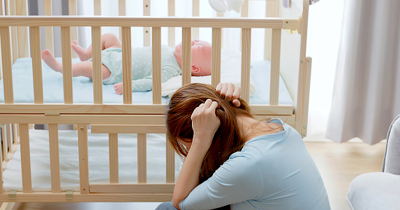 asian mother taking care her baby without patience sitting floor at home has postpartum depression and crying infant is lying on crib