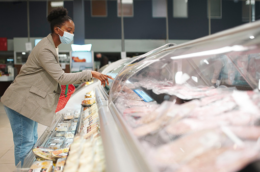 Black woman, customer and supermarket while shopping for groceries during covid with a face mask making choice for cold meat at butcher. Female at retail store for food on display during covid 19