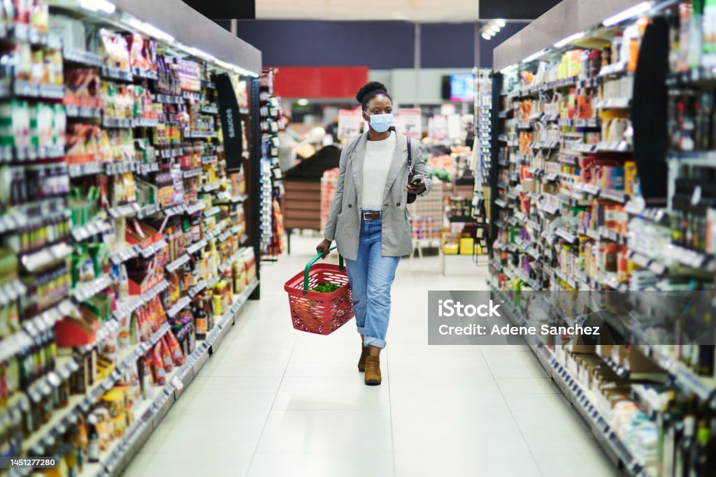 Black woman, face mask and supermarket shopping, covid safety and public health risk in retail shopping mall. Pandemic, market and customer walking in grocery store with basket for products on shelf 25-29 Years Stock Photo