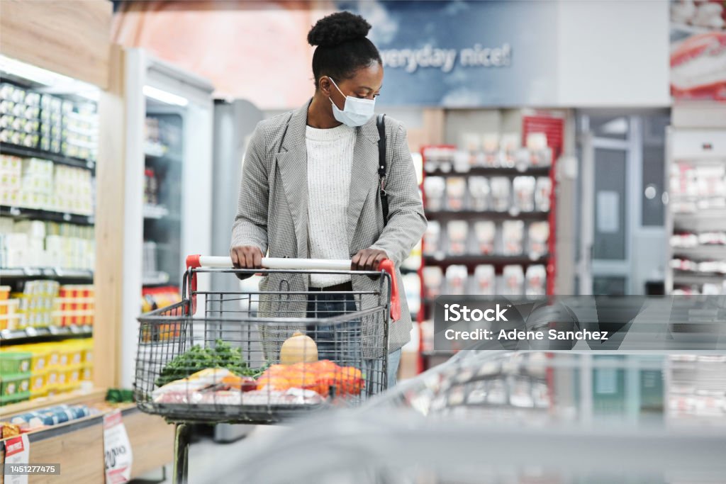 Black woman, shopping cart and supermarket with mask safety, covid or wellness while walking by fridge. Woman, shopping and grocery in retail store with ppe on face by freezer, frozen food or product 25-29 Years Stock Photo
