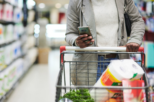 Black woman, hands and phone with trolley for shopping, checklist or ecommerce at the mall. Hand of African American female shopper buying groceries in a store for home essentials holding smartphone
