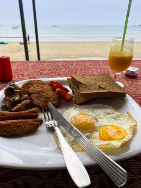 imagem da cena do café da manhã ao ar livre do restaurante da praia, fritar salsichas, ovos fritos ensolarados, fatias de tomate, hash brown, torradas integrais no prato branco, porção única de manteiga e geleia, faca, garfo, copo de suco de laranja,  - orange mushroom asia brown - fotografias e filmes do acervo