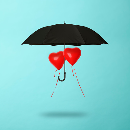 Heart-shaped balloons shared black umbrella floating in mid-air against light blue background.