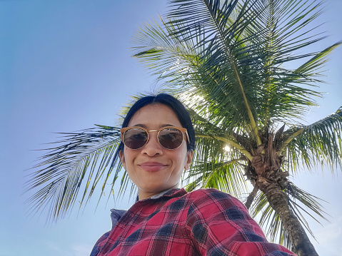 A young Asian woman taking a selfie with a coconut palm tree and blue sky background. Tourist on vacation in Thailand, Southeast Asia.