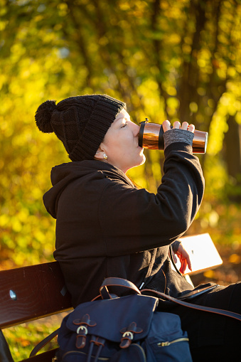 Lady sits on a bench outside and drinks a hot coffee to warm a little bit from the cold autumn temperature. There are colorful leaves of the fall season in the background.