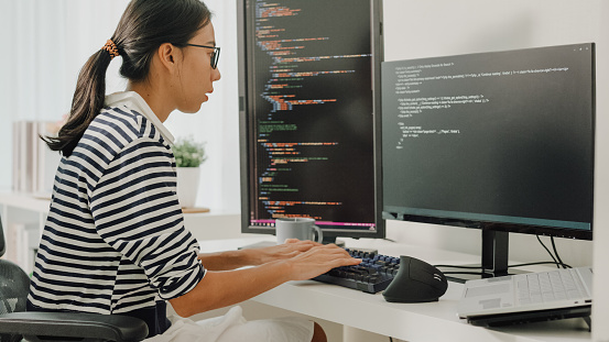 Young Asian woman software developers using computer to write code sitting at desk with multiple screens work remotely at home. Programmer development concept.