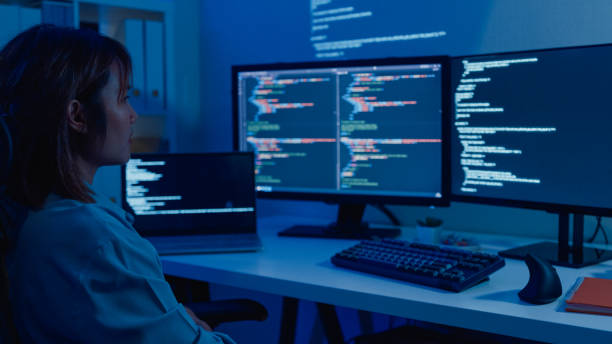 Young Asian woman software developers using computer to write code sitting at desk with multiple screens work remotely in home at night. stock photo