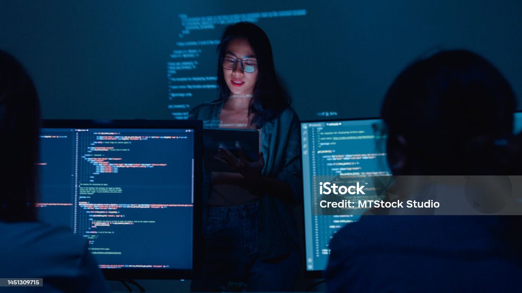 Young Asian woman software developers mentor leader manager talking to executive team analyzing source code in office at night. Young Asian woman software developers mentor leader manager talking to executive team analyzing source code in office at night. Programmer development concept. Technology Stock Photo
