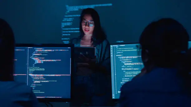 Photo of Young Asian woman software developers mentor leader manager talking to executive team analyzing source code in office at night.
