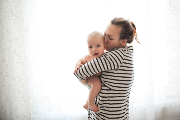 funny baby 6 months mom in her arms against window funny baby 6 months mom in her arms against the window. Mom tickles and gently kisses the baby. Concept maternal love and motherhood, soft focus, toning and lifestyle half dozen babies stock pictures, royalty-free photos & images