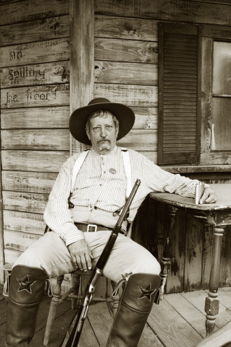 Very somber and tired Caucasian man with sheriff badge sitting on porch holding rifle and flask.  Black and white.   - See lightbox for more