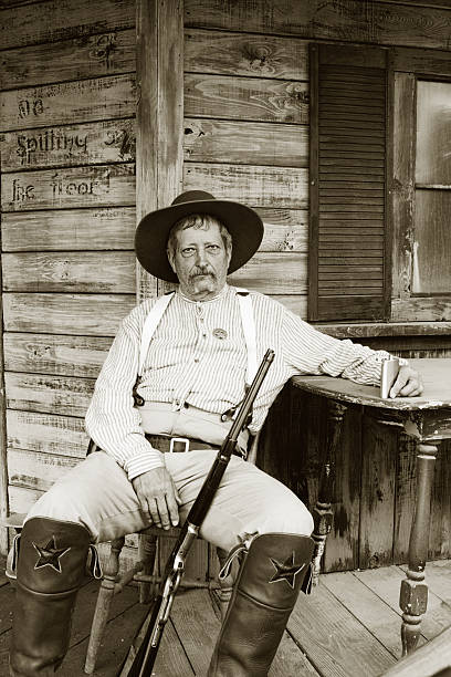 Weary Old Cowboy Sheriff Sitting with Rifle on Weathered Porch Very somber and tired Caucasian man with sheriff badge sitting on porch holding rifle and flask.  Black and white.   - See lightbox for more wild west photos stock pictures, royalty-free photos & images