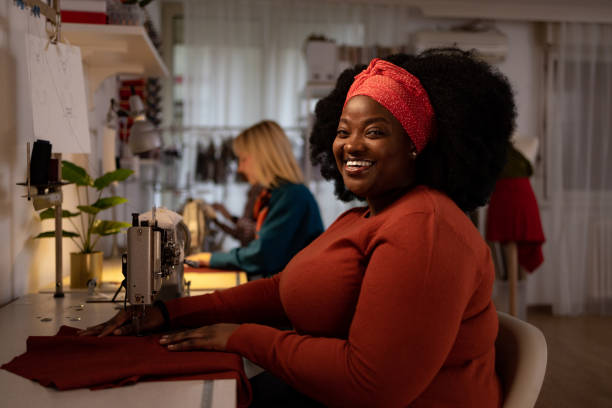 Happy black female tailor enjoying while working on sewing machine in a studio and looking at camera. Her colleagues is in the background.