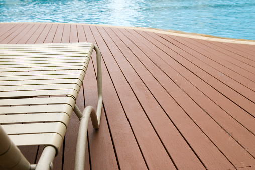 A deck chair on a wooden deck by a swimming pool.