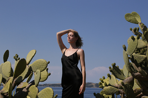 Portrait of young female model standing near big cactuses in black silk satin camisole dress. Stylish and elegent summer fashion.