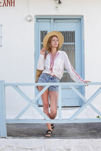 Young fashionable woman in cotton shirt and denim shorts with straw hat posing on the street of Mediterranean seaside village. Summer travel lifestyle concept.