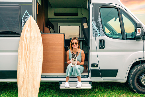Young woman sitting with her boston terrier dog at the door of her camper van during a trip
