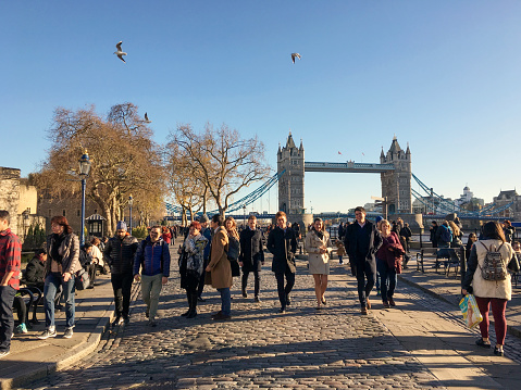 London, UK - Nov 2016: Crowd of people walking on street by river Thames and Tower of London in sunny morning with Tower Bridge and clear blue sky background.