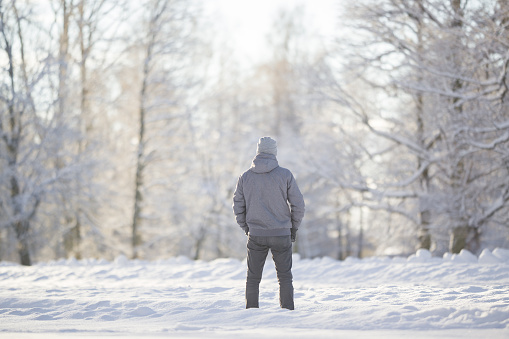 Young adult man standing alone on snowdrift and looking on snow covered trees at park. Spending time alone in nature. Peaceful atmosphere. Back view. White cold snowy sunny winter day.
