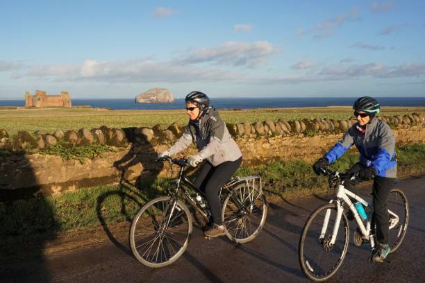 Female cyclists near Tantallon Castle and Bass Rock North Berwick, East Lothian, Scotland - 20 December 2022: A couple of female cyclists are on a country road in East Lothian in the winter sunshine. In the background are Tantallon Castle and the Bass Rock. north berwick scotland east lothian castle scotland stock pictures, royalty-free photos & images