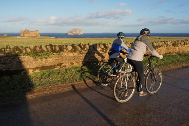 Female cyclists near Tantallon Castle and Bass Rock North Berwick, East Lothian, Scotland - 20 December 2022: A couple of female cyclists are on a country road in East Lothian in the winter sunshine. In the background are Tantallon Castle and the Bass Rock. north berwick scotland east lothian castle scotland stock pictures, royalty-free photos & images