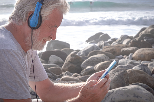 Senior man white beard listening to music with mobile phone wearing headphones and smiling. In background ocean waves and surfers