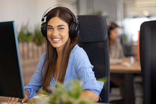 Happy businesswoman having headphones while working on desktop PC at corporate office and looking at camera.