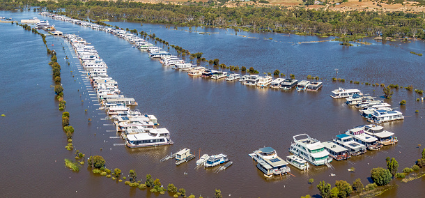 Aerial view flooded Mannum Waters Marina on River Murray, with moored houseboats, submerged access road, surrounding lagoon. River Murray in background between trees. Business disaster during summer holiday period. Panoramic format