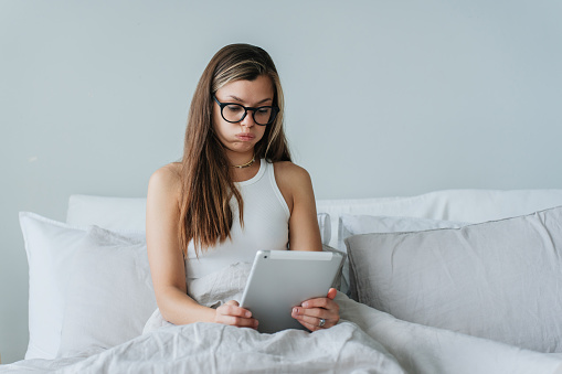 Perplexed young Hispanic woman sitting on bed awakes holds tablet looks at screen puffs out cheeks wears glasses. Pensive caucasian girl checks her overloaded agenda. Tired student.