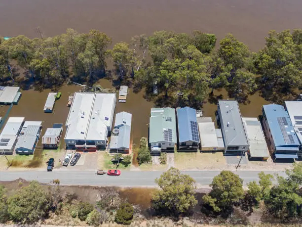 Aerial view, advancing floodwaters inundating shacks and houses at Bolto on the River Murray; red car with trailer on dry part of Khartoum Road. Levee in foreground, flooded trees and riverbank in centre frame with muddy river waters: threats, inundation, ominous. Solar panels and TV antennae, people packing vehicles with belongings.