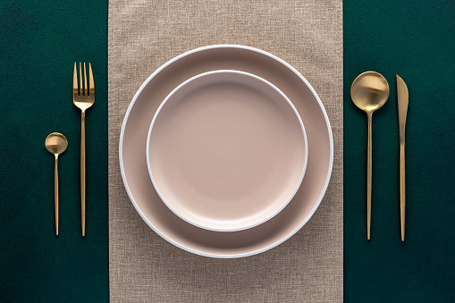 Festive place setting with beige napkin. Empty plates and gold cutlery on dark green background. Top view. Dining table in luxury restaurant. Card or menu template, flat design. Tableware, crockery