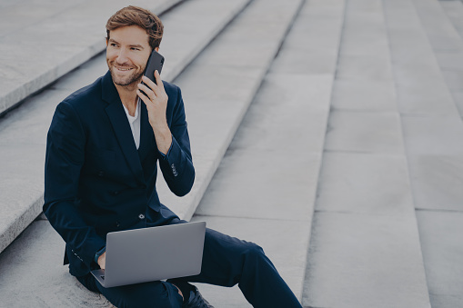 Caucasian satisfied man in formal outfit sitting on steps outdoor, talking on mobile phone and working on laptop computer. Male freelancer typing on keyboard while speaking on cellphone with client