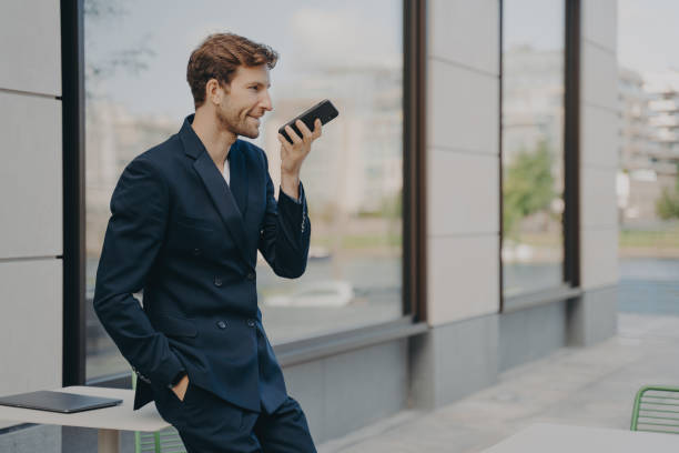 uomo d'affari fiducioso con smartphone che utilizza l'app di assistente vocale mentre si appoggia al tavolo del caffè all'aperto - cafe table outdoors speaker foto e immagini stock