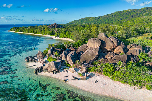 A scenic view of a tropical beach on Similan islands with white sand and clear blue water. On the right side of the image, there are large dark grey boulders covered with dense green vegetation. The sky is blue with white clouds. The image is taken from a low angle, looking up at the boulders and greenery.