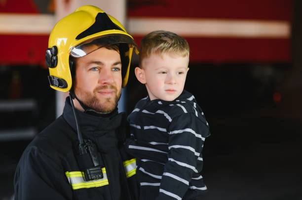 istock Dirty firefighter in uniform holding little saved boy standing on black background. 1451387378