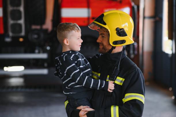 istock Dirty firefighter in uniform holding little saved boy standing on black background. 1451387442