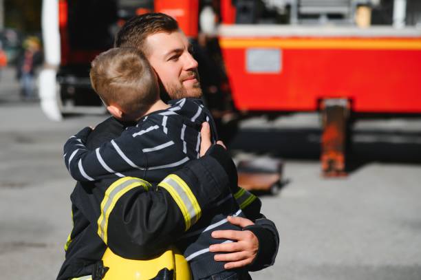 Dirty firefighter in uniform holding little saved boy standing on black background