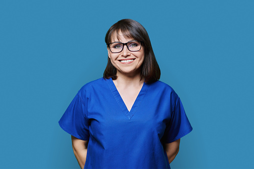 Smiling woman wearing blue scrubs uniform looking at camera on blue color background. Female doctor nurse laboratory assistant pharmacist veterinarian medical worker