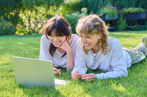 Young teenage female student friends lying on grass with laptop. Teenagers rejoicing laughing and having fun together. Friendship, youth, technology, high school, college, lifestyle concept