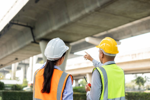 istock Two Asian worker engineer man and woman architect looking construction with white safety helmet in construction site. Standing at highway concrete road site. Work planning with blueprint and tablet. 1451398201