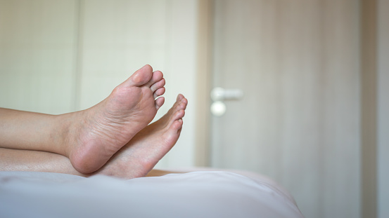 Human legs part during laying or sleeping down on the bed sheet with domestic room interior background. Holiday relaxation concept photo scene, selective focus.