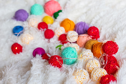 A lot of colorful knitting yarn and a luminous garland in the form of balls lie on a white fluffy blanket. Cozy still life made of yarn. Close up. Copy space