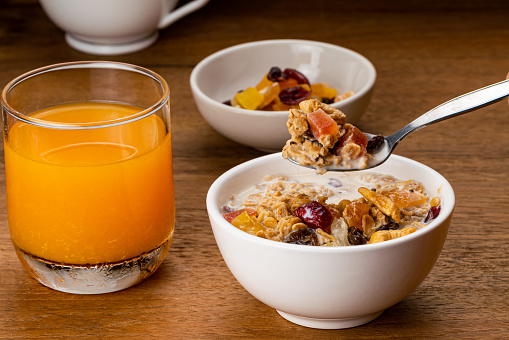 Taking a spoon full of muesli cereal breakfast with fresh milk and preserved sweet tropical fruit in white ceramic bowl with cold orange juice in transparent glass on wooden table.